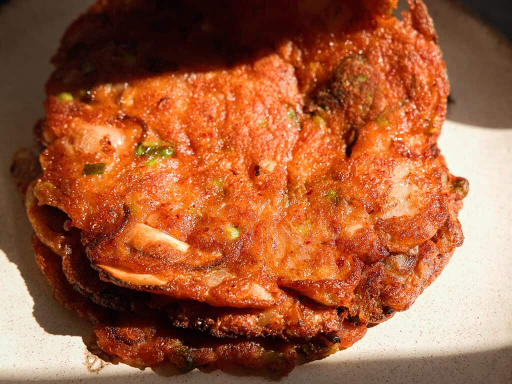 A close-up of crispy, golden-brown Korean kimchi pancakes stacked on a plate, with visible bits of cabbage and green onions, lit by natural sunlight.